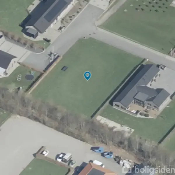 Luftfoto af en parcelhusområde viser en tom grund markeret med en blå nål. Omgivet af huse, veje og græsplæner. Ingen tekst.