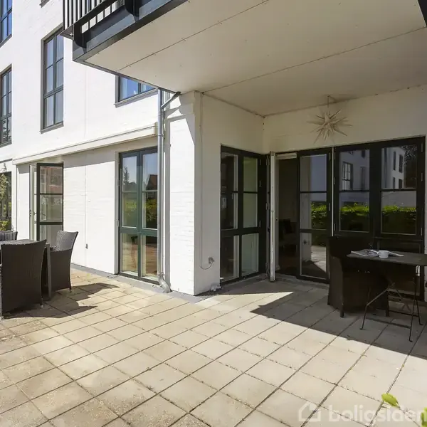 En udendørs terrasse med et bord og seks stole står foran en moderne, hvid bygning med store vinduer. Et skilt viser "11D". Omkring er der grønne buske og blå himmel.