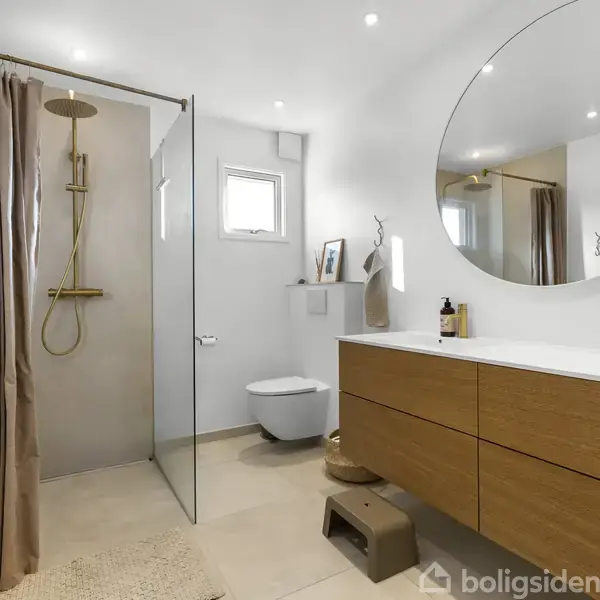 Badeværelse med bruser og glasvæg, træmøbler med hvid bordplade, rundt spejl på væggen, toilet og lille vindue. En krukke og genstande ses som dekoration.