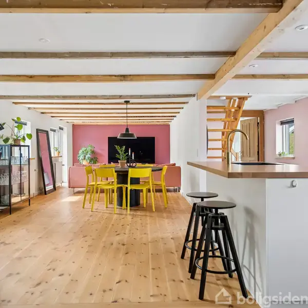 En åben stue med træloft og gulv; en hvid køkkenø med barstole til højre; et spisebord med gule stole i midten; planter og lysindfald fra vinduer.