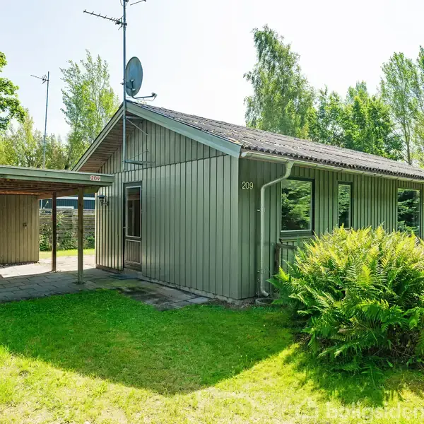 Et grågrønt hus med nummeret 203 står i en solbelyst have med frodigt grønt græs og buske. En carport ved siden af huset står på fliser, omgivet af træer.