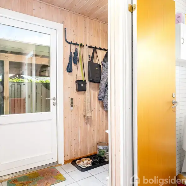 Hvid dør med vindue åbner til entré med trævægge; kroge med tasker og jakker på væggen. Til højre er en dør til et lille badeværelse med toilet.