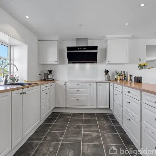 Et moderne køkken med hvide skabe og træbordplader. En stor emhætte hænger over komfuret. Køkkenet er lyst op af vinduer med udsigt til en grøn have.