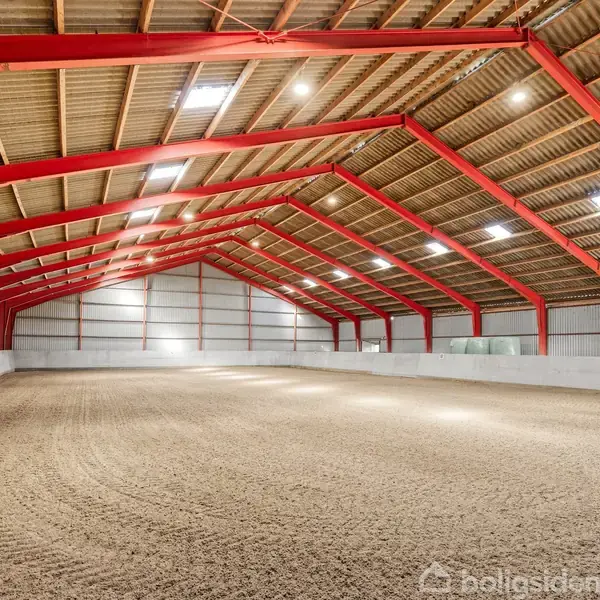 En stor indendørs ridehal med rødmalet stålstruktur, sanddækket gulv og træloft. Hallen er oplyst af loftslamper og naturligt lys fra ovenlysvinduer.