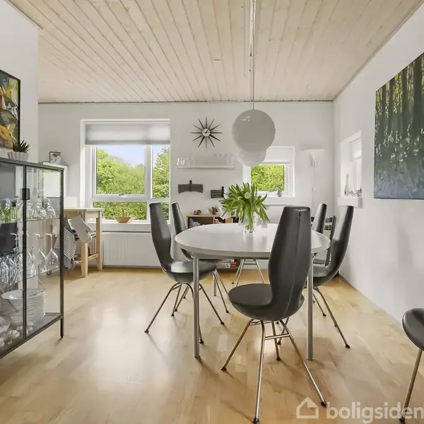 Et spisebord med sorte stole omgives af et lyst rum. Et glasvitrineskab står til venstre med porcelæn og glas. Store vinduer lader naturligt lys komme ind, og moderne kunst hænger på væggene.
