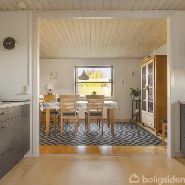 Spisestue med et spisebord med seks stole, omgivet af hvide vægge og træloft. Et stort vindue giver naturligt lys. Køkkenartikler ses i forgrunden.