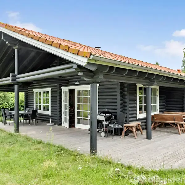Et sort træhus står på en træterrasse med bord og grill, omgivet af grøn græsplæne. Flere hvide døre og vinduer pryder facaden, mens græs og træer omkranser området.