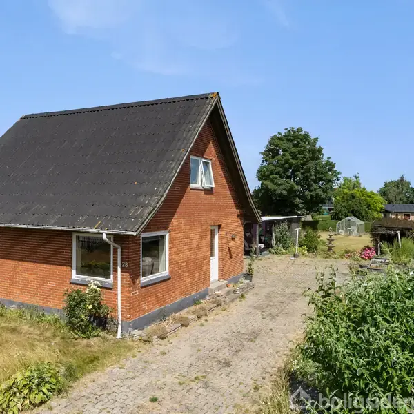 Rødt murstenshus med sort tag står på en brolagt indkørsel. I baggrunden ses have med græs, buske og træer under en skyfri himmel.