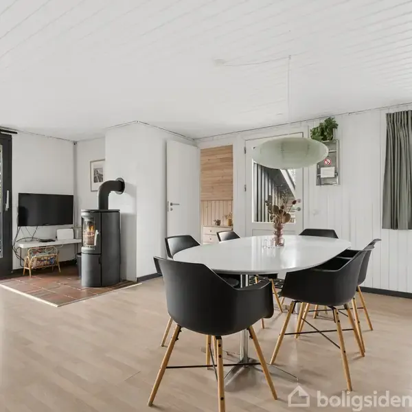 Et lyst stueområde med et rundt spisebord omgivet af fire sorte stole, en brændeovn ved en væg, og et TV i hjørnet. Store vinduer giver udsigt til omgivelserne udenfor.
