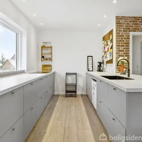 Gråt køkken med hvide bordplader. Handlingsfri scene. Kontekst inkluderer murstensvæg, opslagstavle, stor vask, vindue med udsigt og træhylde med få genstande. Flot træparketgulv lyses op af spotlys.