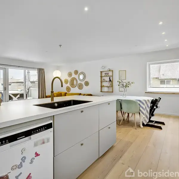 Køkken med stor hvid ø med vask, omgivet af spisebord og stue med sofa, dekoreret med vægkunst og blomster, belyst af naturligt lys fra vinduer.