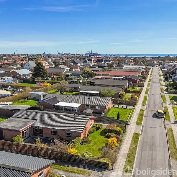 Række af enfamiliehuse står stille langs en asfalteret vej i et forstadsområde. Omkring husene er der grønne haver, og i baggrunden ses skibe ved en havn under en klar blå himmel.