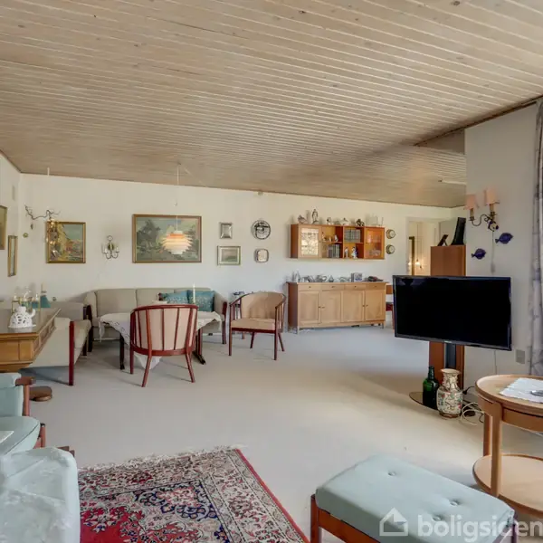 Stue med lys sofa og stole om et sofabord, fladskærms-TV på stander; lyse vægge og loft; reoler med pynt og bøger i baggrunden. Store vinduer med gardiner til højre.