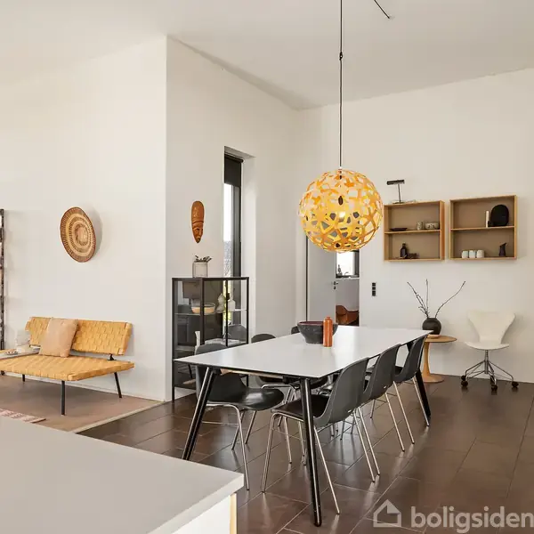 En moderne spisestue med et langt bord og sorte stole. En rund, gul lampe hænger over bordet. Rummet er lyst med minimalistisk indretning og få dekorationer på væggene.