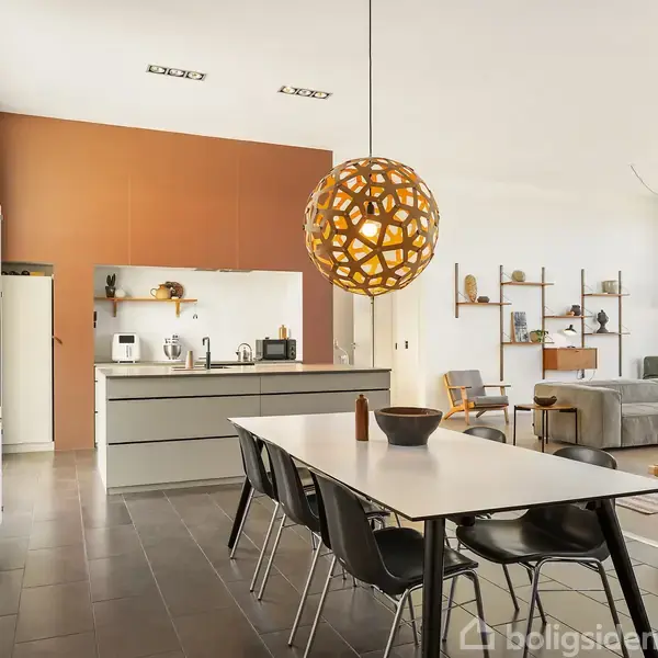 Spisebord står under en dekorativ lampe i et moderne, åbent køkken og stueområde. Køkkenet har grå skabe og apparater, mens stuen har sofa og vægmonterede hylder.