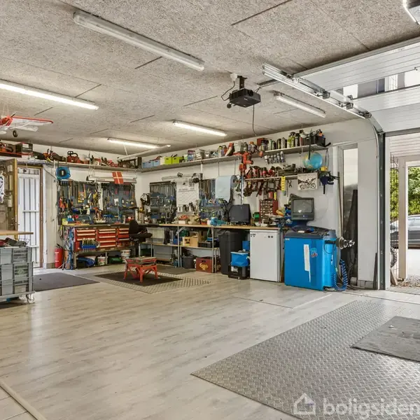 Et veludstyret værksted med værktøjer hængende på væggene og borde fyldt med udstyr. Åben garageport afslører bil parkeret udenfor, træer i baggrunden. Ingen tekst fundet.