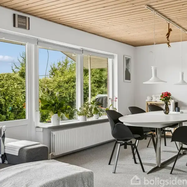 Spisebord omgivet af seks sorte stole står i en lys stue med store vinduer, der viser udsigt til en frodig have. En sofa og billeder dekorerer rummet.