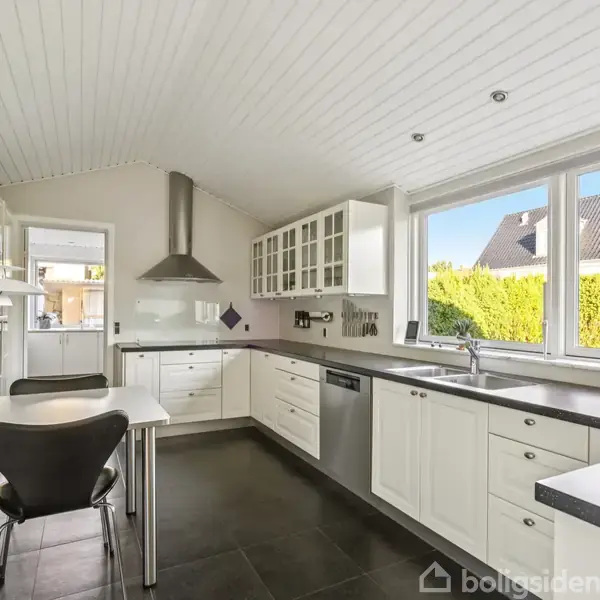Hvidt køkken med sorte gulvfliser, moderne apparater, og spisebord med sorte stole. Store vinduer giver udsigt til en have med hække og et nærliggende hus.