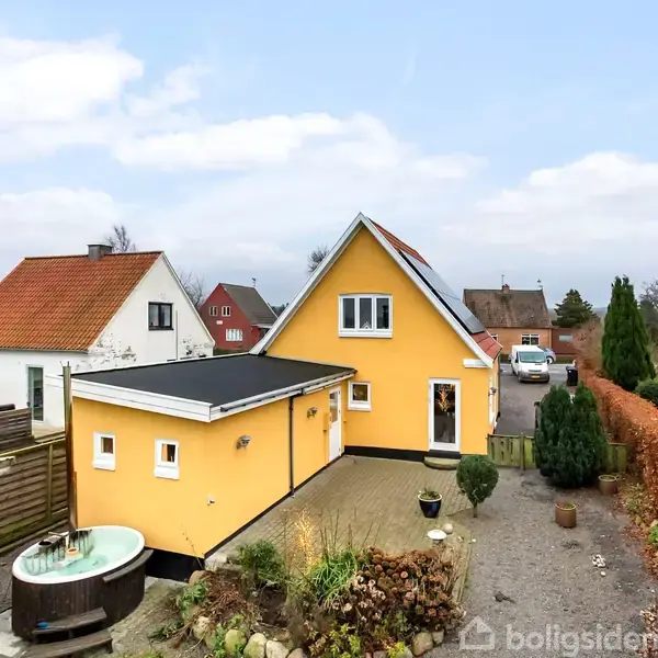 En gulmalet villa med spidst tag står i en forstadshave. Huset har en terrasse og et spabad i nærheden, omkranset af hegn og andre villaer.
