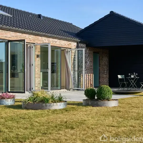 Et moderne hus med glasdøre står åbent mod en terrasse. En lille gårdhave med bord, stole og planter er omgivet af en velplejet græsplæne.