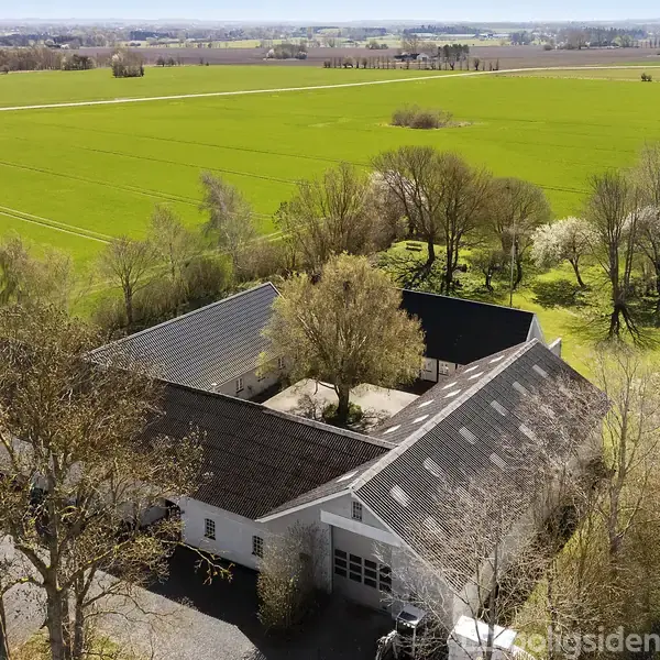 En gård med hvid facade og sort tag omkranser et gårdhaverum med et stort træ i midten. Omgivet af grønne marker og bevoksede træer i landligt område.