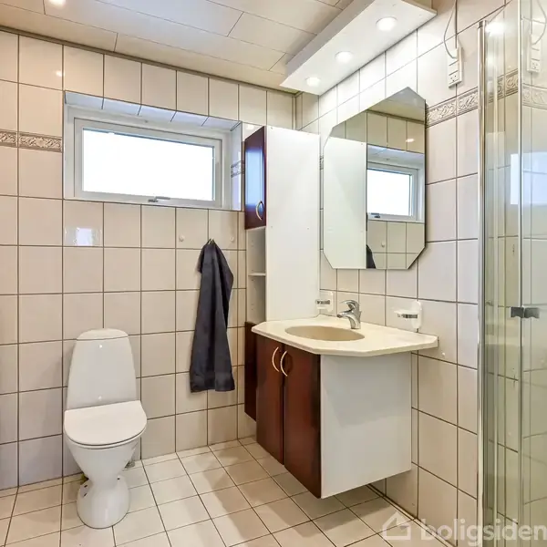 Toilet placeret ved siden af et håndklædetørrer, omgivet af flisebelagte vægge. En vask med skab og spejl hænger ved siden af en glasbrusekabine. Enkelt hvidt vindue i baggrunden.