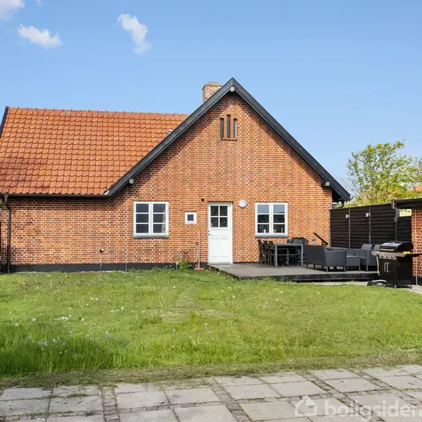 Rødt murstenshus med skråt orange tag står foran en grøn græsplæne. En terrasse med sorte havemøbler er placeret ved husets indgang. Omgivet af træer under en klar blå himmel.