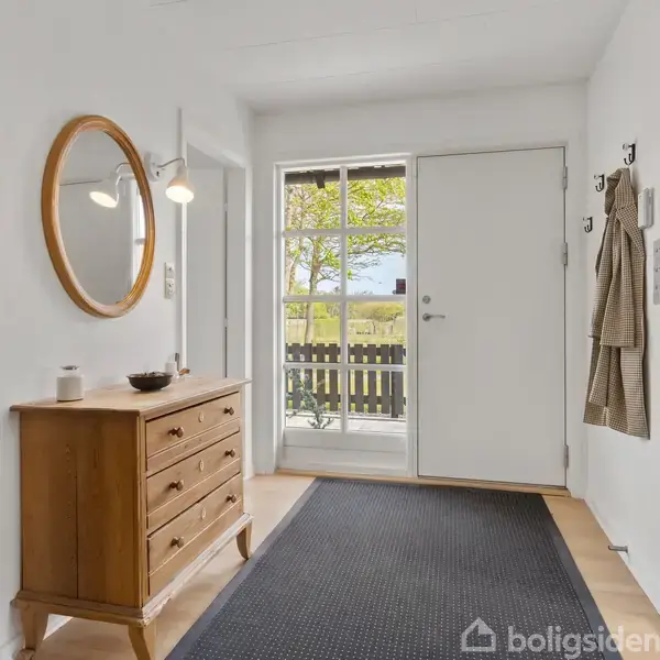 Et træskommode står i en lys entre, dekoreret med lys og spejl, ved siden af en dør med vindue, der viser udsigt til en grøn have.