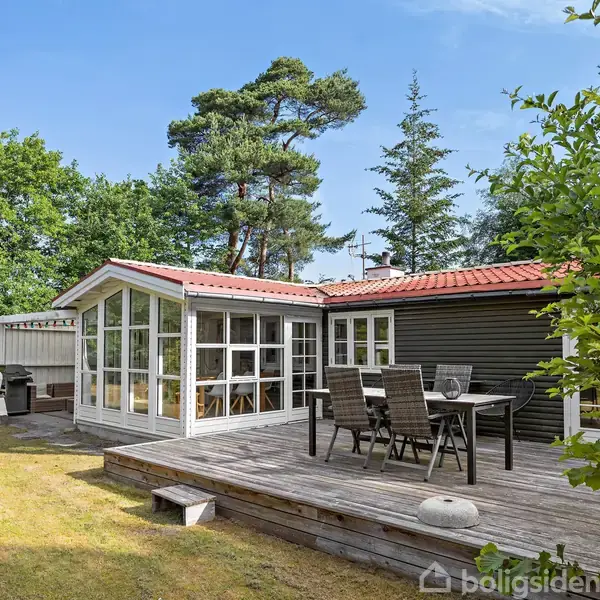 Sommerhus med træbeklædning og rødt tag står på en terrasse med havemøbler. En udendørs pejs er placeret til venstre. Omgivet af træer og grønne områder.