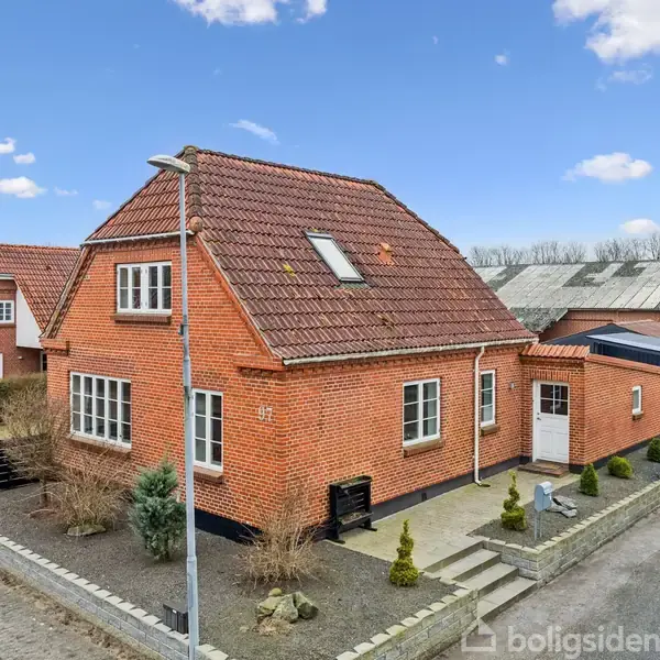 En rød murstensvilla med rødt tegltag står på en hjørnegrund, omgivet af en flisebelagt gangsti og små buske. Huset har flere vinduer og en synlig dør.