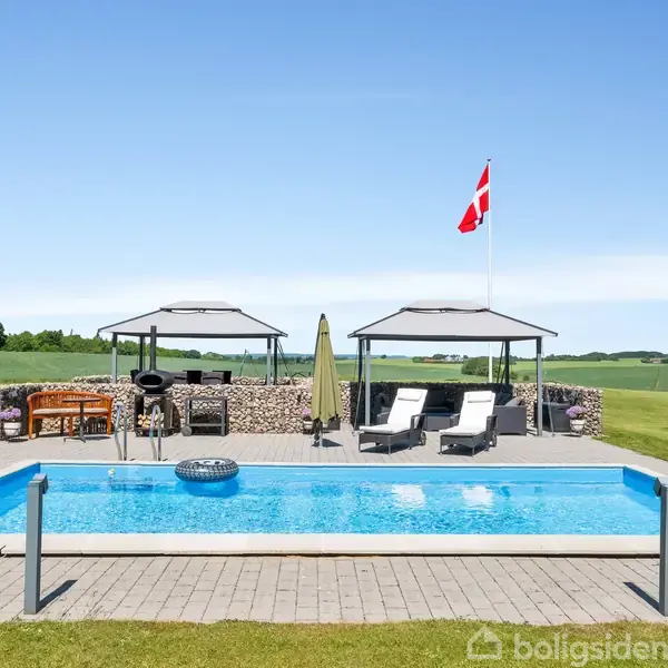 En swimmingpool omgivet af en flisebelagt terrasse med solsenge og parasoller i en grøn have. Dannebrog vajer på en flagstang i baggrunden under en klar blå himmel.