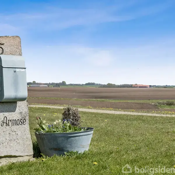 En stenblok med en postkasse mærket "5 Ø Årmose" står på en græsplæne. En plantekrukke er foran, omgivet af fladt landskab med marker i baggrunden.