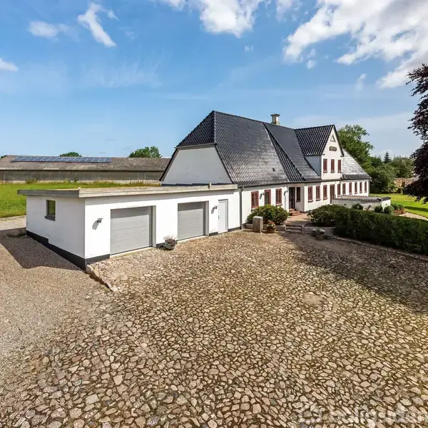 Hvid hus med sort tag står i en gårdsplads omgivet af åbne marker og træer, under en klar blå himmel med lette skyer. Der ses flere garager og velplejet græsplæne.