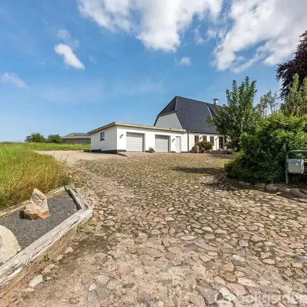 Et hvidt hus med mørkt tag står stille for enden af en stenbelagt indkørsel. Det er omgivet af grønne områder og en blå himmel med skyer. To postkasser ses til højre.