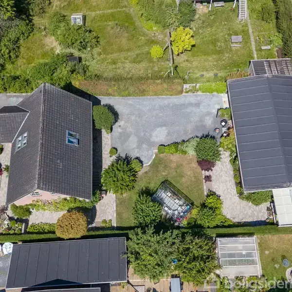To huse med mørke tage står ved siden af hinanden, adskilt af en grusplads. Området er omgivet af grønne planter og en vej løber til venstre for ejendommene.
