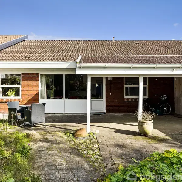 En murstenshus med rødt tag står i et solbeskinnet område. En flisebelagt terrasse har havemøbler og planter. Til højre ses en cykel opbevaret under et halvtag.