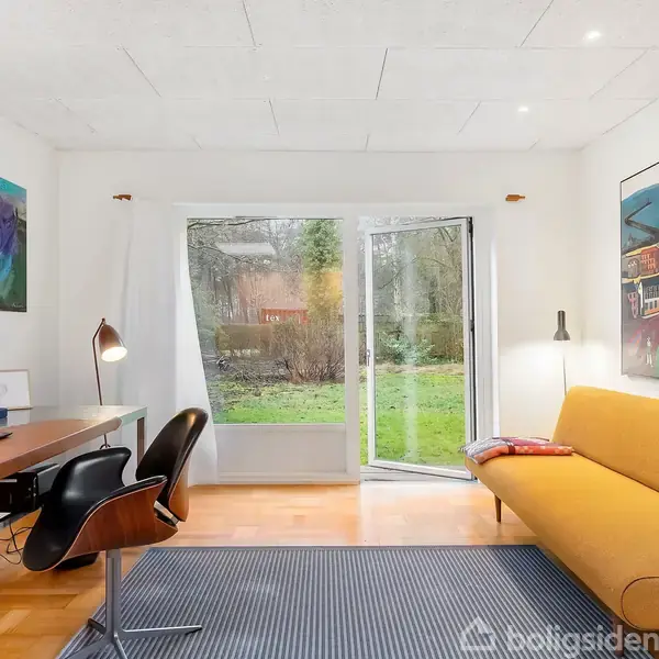 Gult sofamøbel står ved siden af et skrivebord med en lampe og laptop; begge vender mod en stor vinduesdør til en grøn have. To farverige malerier hænger på væggene.