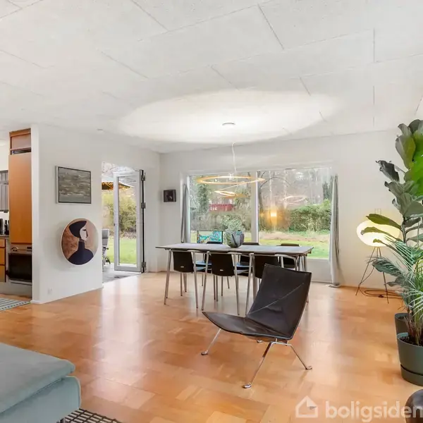 En lys stue med spisebord og stole står foran store vinduer, der åbner op til en grøn have. En stol og en stor plante er til venstre.