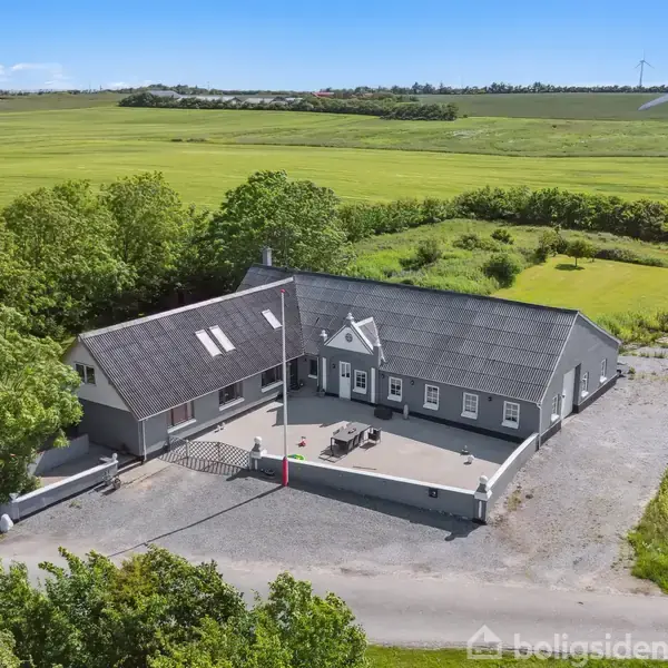Grå bygning med sort tag og flere vinduer omgivet af grusareal. Bagved bygningen ses grønne marker og en vindmølle på en solskinsdag.