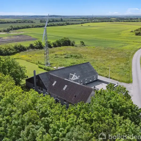 En stor, mørkt tagbeklædt bygning ligger blandt grønne træer. En snoet landevej ses ved siden af, omgivet af vidstrakte marker. Baggrunden viser en næsten skyfri himmel.