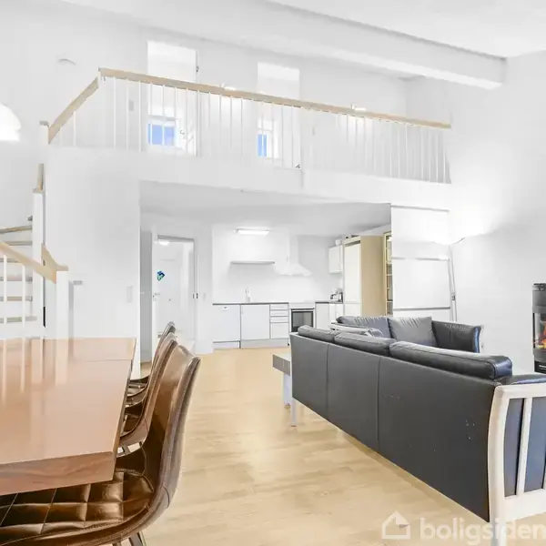 En moderne stue med et sort sofaarrangement omkring en brændeovn, står i et rum med åbent loft, køkken i baggrunden og en trappe til en overetage, indrettet med lyse møbler.