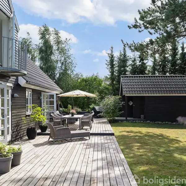 En træterrasse med havemøbler og en parasol strækker sig fra et sort, træklædt hus. Der er grønne planter omkring, og en græsplæne med et skur i baggrunden.