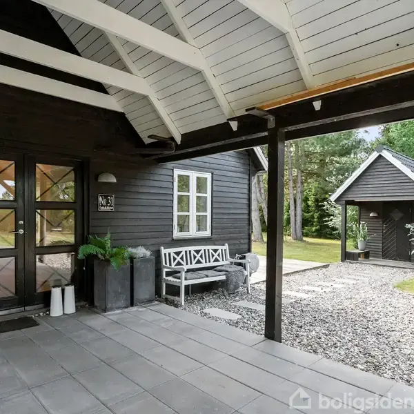 Sort træhus med glasdør og bænk foran. Flisebelagt terrasse med udsigt over grusbelagt gangsti. Omkranset af træer og græsplæne.