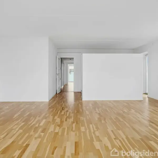 Stue med lakeret trægulv, hvide vægge og loft. Flere døråbninger leder til andre rum. Tre store vinduer slipper lys ind. En radiator er synlig til højre.