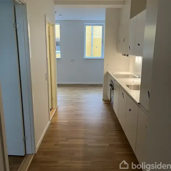 Et smalt køkken med hvide skabe langs højre side. To vinduer i baggrunden giver lys. Trægulv strækker sig gennem rummet mod en åben dør i venstre side.