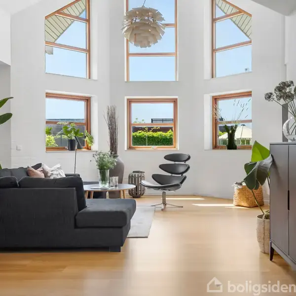 Stue med moderne sort sofa og lænestol, store vinduer lader masser af lys ind, lyse trægulve og grønne planter omkring.