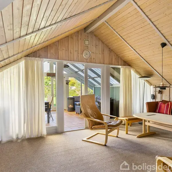 Et hyggeligt loftsrum med træbeklædte vægge har en lænestol og sofa ved et sofabord. Gennemsigtige gardiner hænger foran glasdøre, der åbner til en terrasse med møbler.