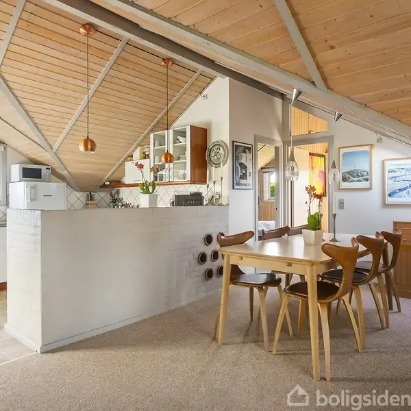 Et lyst køkken-alrum med træloft og skråvægge. Et spisebord med træstole står i rummet. Køkkenet har åbne hylder og en bardisk. Vægge dekoreret med malerier.