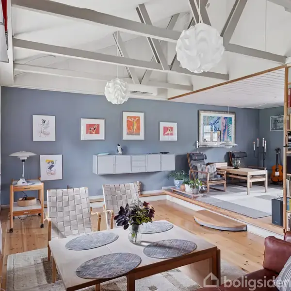 Stue med sofabord dækket med dækkeservietter, omgivet af læder- og fletmøbler. Vægge dekoreret med kunstværker. Reol fyldt med bøger. Lysekrone-lys hænger fra loftet.