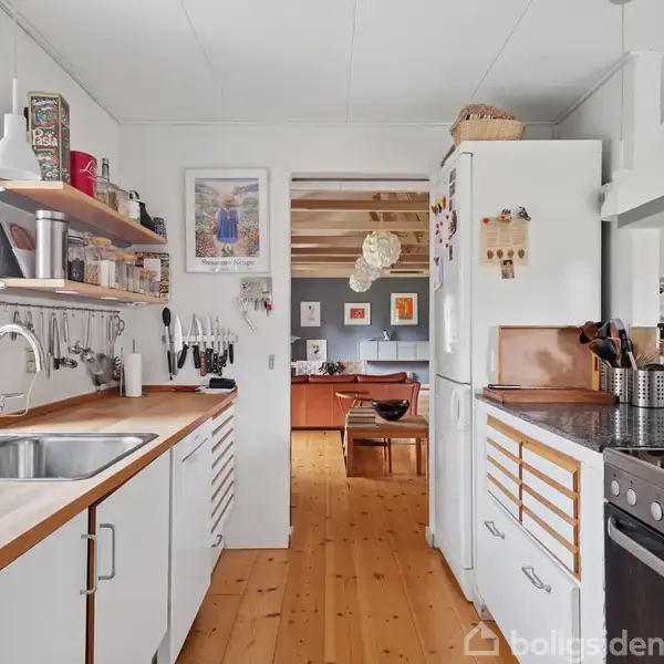 Et moderne køkken med hvide skabe, en vask og ovn. Køkkenet fører ind til en stue gennem en åben døråbning. Trægulve og vægdekorationer skaber en varm atmosfære.